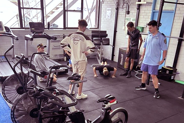 IPROWD students in a gym complete fitness training with NSW Police. One student is doing a push up, another is on their knees. Three others are standing. There are treadmills, weight machines and bikes on the floor.