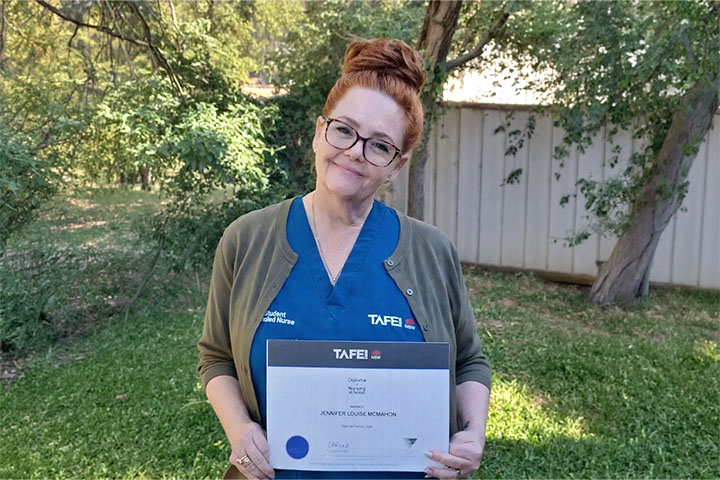 Jennifer McMahon stands in a sundrenched, fenced garden. She wears a TAFE NSW blue nursing uniform and smiles proudly at the camera while holding her Diploma. Jennifer red hair is tied in a neat knot on her head. She wears tortoise-shell rimmed glasses.