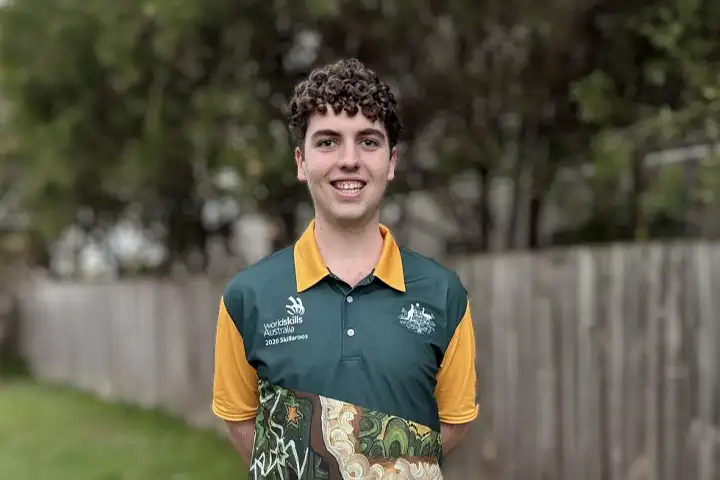 John Ray stands outside with grass, trees and a wooden fence in the background. He’s looking directly at the camera with a big smile. He wears a green and yellow WorldSkills Australia polo shirt.