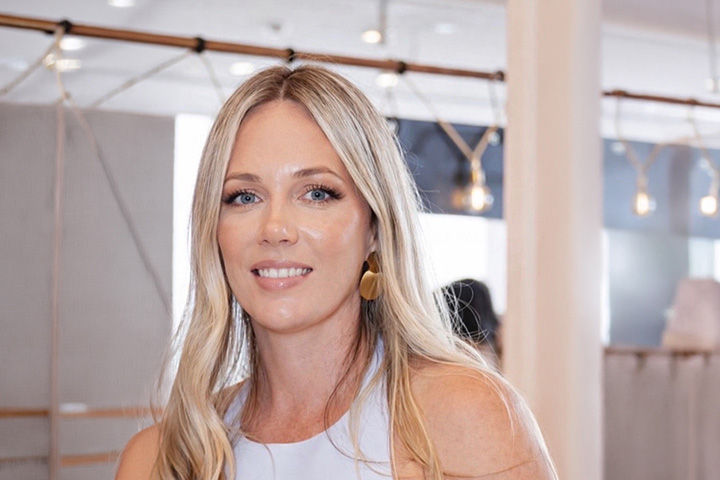 Kat Gray smiles happily at the camera. She’s inside a well-lit, white room with activity in the background. She wears a white sleeveless top and has long light-coloured hair.