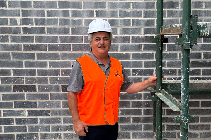 Illawarra builder, Ken Tugrul, from TDK Corporation stands in front of a newly laid grey brick wall. He wears a white hard hat and an orange hi-vis safety vest. Ken is smiling directly at the camera. His grey hair is visible on either side of his hard hat.  