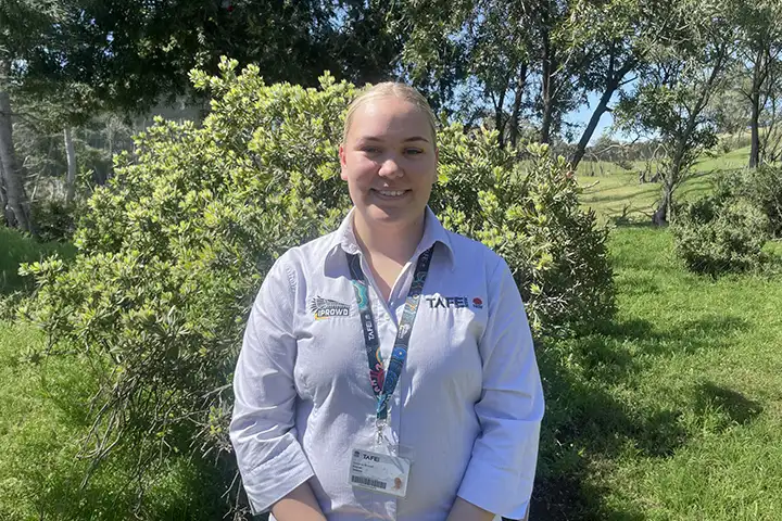 Kolarah Newman, a proud Wiradjuri and Wailan woman from Dubbo, stands in a garden at her IPROWD graduation. She smiles proudly at the camera. Her hair is tied back off her face, and she wears a TAFE NSW IPROWD long-sleeved shirt.