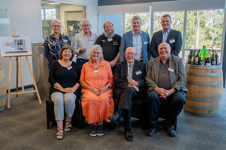 The 9 founders at TAFE NSW Kurri Kurri Winery are positioned in two rows, smiling proudly. To their right is a wine barrel holding opened wine bottles and to their right stands an easel.