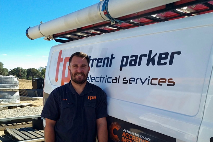 Lachlan James stands outside leaning against his work van. He’s smiling directly at the camera wearing a short-sleeved, dark blue, collared work shirt. Lachlan has brown hair, a beard and moustache. There are stacked building materials in the background. It is a clear, sunny day.