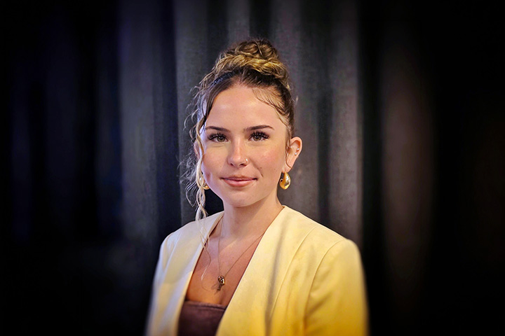 TAFE NSW Bathurst graduate Laci Petford wears a yellow blazer over a purple top. She is smiling and standing confidently at the camera against a black curtain background.