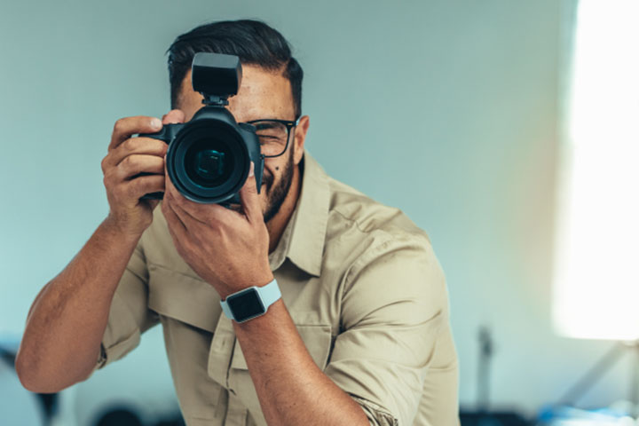 Man wearing beige button shirt with rolled sleeves looks straight ahead with a camera held up to his face.