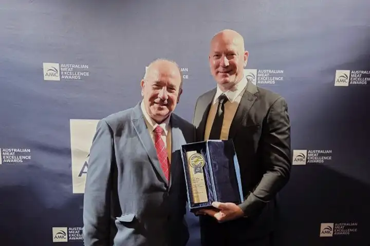 Martin Paynter, TAFE NSW Granville Head Teacher of Meat and Allied Trades and TAFE NSW Team Lead stand holding their AMIC Best Overall award. They’re wearing business suits, white shirts and ties standing in front of a dark blue, branded award curtain.