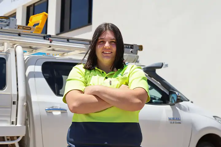 TAFE NSW electrical apprentice Molly stands proud, arms crossed in a hi-vis shirt. Behind her is a white work vehicle.