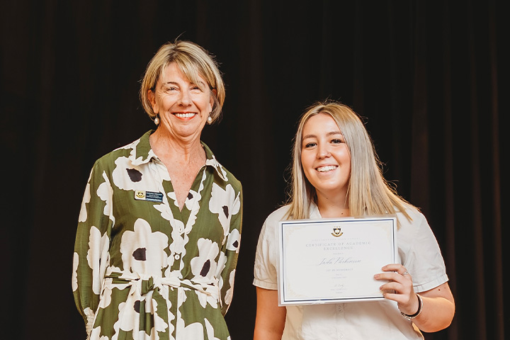 Jada and her teacher are standing smiling in front of a black back drop. Jada is holding her certificate up proudly.