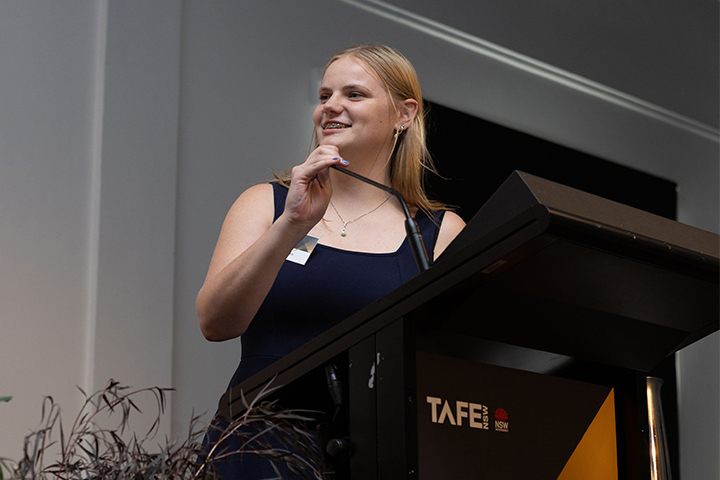 Ruby Beckenham is standing at a podium in a navy gown delivering her speech on behalf of all award recipients at the 2026 TAFE NSW Student of the Year Awards. 