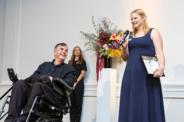 Ruby Beckenham is standing on stage in a navy gown delivering her speech on behalf of all award recipients at the 2026 TAFE NSW Student of the Year Awards. She stands in front of a bright bouquet of flowers and next to a gentleman sitting in a mobility device, smiling up at her.
