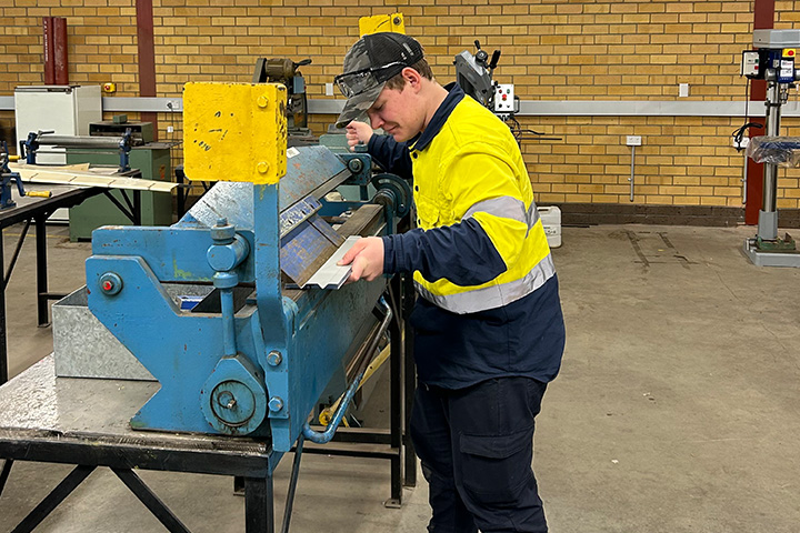 A man wearing a high-visibility yellow and navy work shirt, safety glasses, and a cap operates a large sheet metal bending machine in a workshop. 