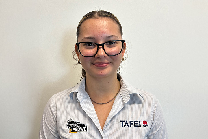 IPROWD graduate Shyama Farrelly looks directly at the camera, smiling. She’s wearing a white TAFE shirt, silver necklace and black glasses with her hair pulled back. She stands against a plain white background. 