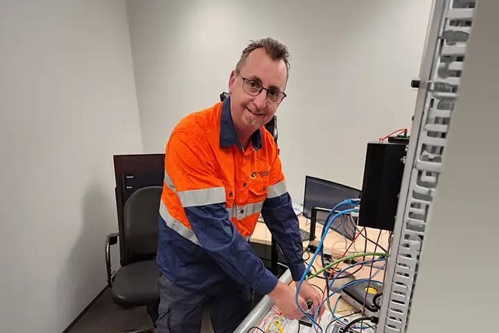 Steven Ellis is working with several coloured wires in an office setting. He’s looking up from his work, smiling at the camera. He wears a collared orange and navy blue high-vis work shirt and metal rimmed glasses. 