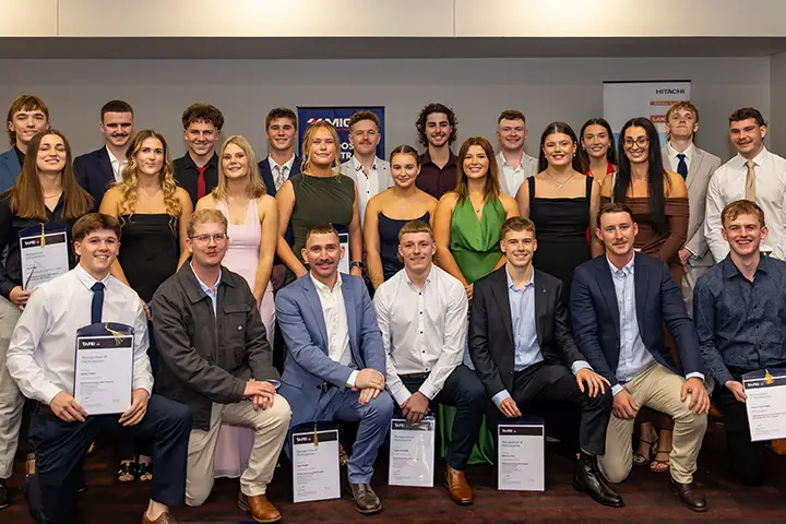 Muswellbrook Mining and Energy Skills Program graduates pose for a photo with their certificates. There are 3 rows of students, the front row kneeling on one knee. The graduates are dressed in suits and dresses, and are all smiling at the camera. 