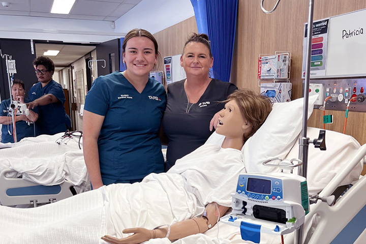 A smiling Talia Opetaia stands  beside her teacher in a simulated hospital setting. She wears a blue short-sleeved uniform with her hair tied back off her face. A dummy patient lies in a bed attached to a heart monitor machine. 