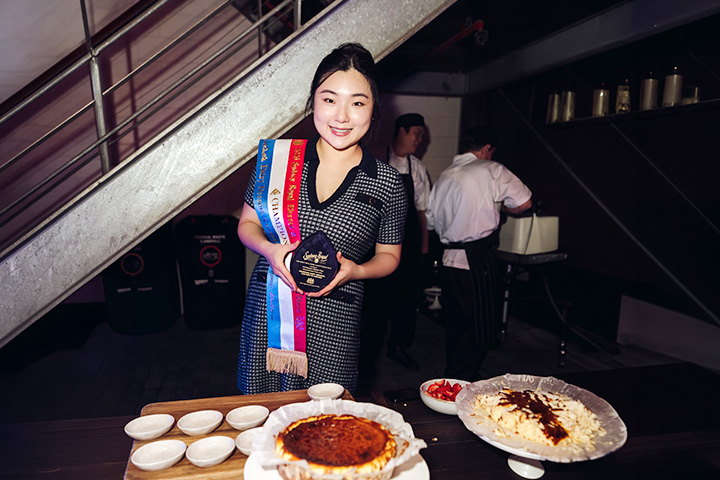 Vivian Wang stands smiling behind a table with desserts. She holds her Sydney Royal National Champion award and a winner’s sash with gold tassels is draped over her right shoulder.