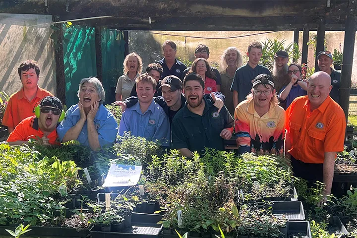 TAFE NSW graduates from the Byron Bay Herb Nursery’s disability support program stand in a light-filled outdoor greenhouse. They are smiling at the camera. People in the front row are leaning on a table filled with seedlings in trays.