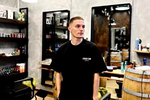 Zeke Smith standing inside the barber shop. He is wearing a black Leisure Club t-shirt, hand in his pockets looking away from the camera.  