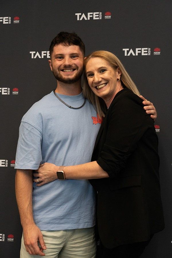 Caleb Sturges, a 20 year-old nursing graduate at TAFE NSW Kingswood, smiles happily. There is a TAFE NSW banner board in the background. 