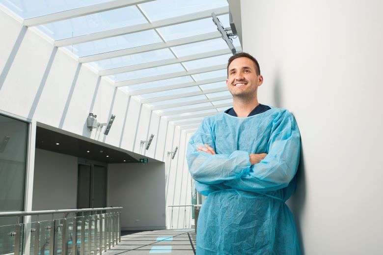 A young man wearing a nurse’s uniform of blue scrubs smiles confidently. He has his arms crossed while leaning against a wall in a hospital or medical centre.