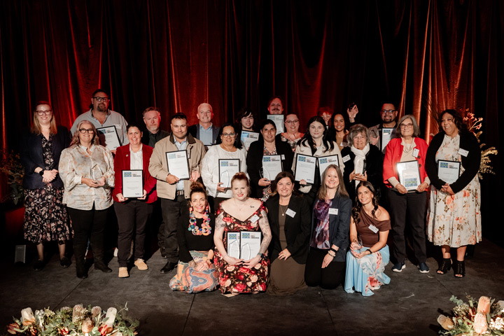 A large group of people, appearing to be recipients of the 2026 TAFE NSW Gili Awards, are posing for a photograph on a stage in front of a dark red curtain. Many are holding framed certificates, and some are holding glass trophies. The lighting is warm, and a floral arrangement is visible in the foreground.