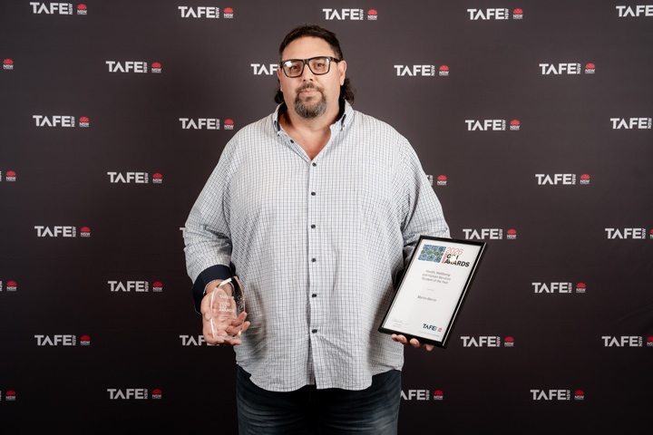 Marty Morris holds his 2026 Gili Award trophy and certificate, one in each hand. He stands in front of a black TAFE NSW backdrop, wearing black glasses and a light checked shirt. 