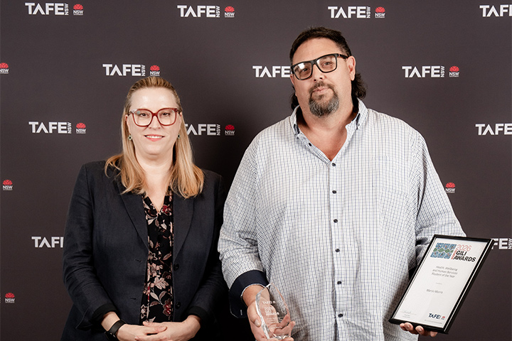 Marty Morris holds his 2026 Gili Award trophy and certificate, one in each hand. He stands next to Chloe Read, TAFE NSW Managing Director. They are both smiling and looking at the camera against a black TAFE NSW backdrop.
