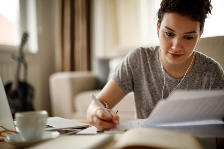 A woman sits at her desk with a tea, her headphones in and pen to paper. She looks content as she looks at a document and has her laptop and notebook open on her desk.