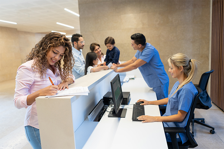 In a bright and modern reception area, a woman with curly hair is smiling as she fills out a form at a counter. Behind the counter, a female receptionist in medical scrubs is working on a computer. In the background, a healthcare professional in scrubs is talking to a family with two young children, creating a warm and welcoming atmosphere.