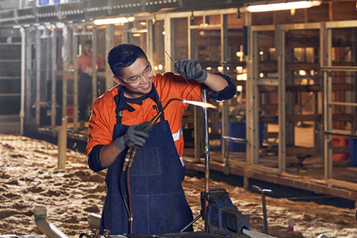 A tradesman working at night with specialised equipment and tools. Other workers are in the background, kneeling and working. The outdoor space has a semi-constructed building lit up with strong outdoor lights.  