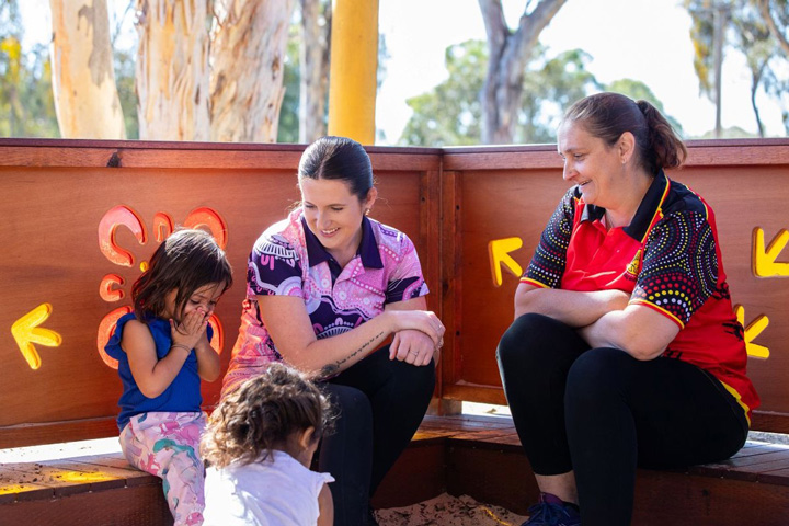 Early childhood education students sit with young children in an outdoor play area, engaging in hands‑on learning with sand and natural materials inside a wooden play structure, with trees and playground features visible in the background.