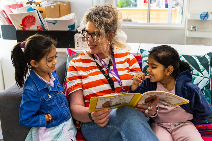TAFE NSW early childhood educator sits on a couch reading a children’s book with two young children, holding the book open while the children look on, with colourful cushions, artwork, and classroom materials arranged in a bright learning environment.