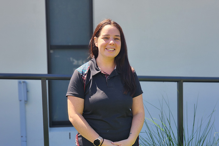 Participant in the ECEC Professional Learning Program stands outdoors in front of a modern building, wearing a black polo shirt and smartwatch, with a railing and landscaped greenery visible in the background under bright daylight.
