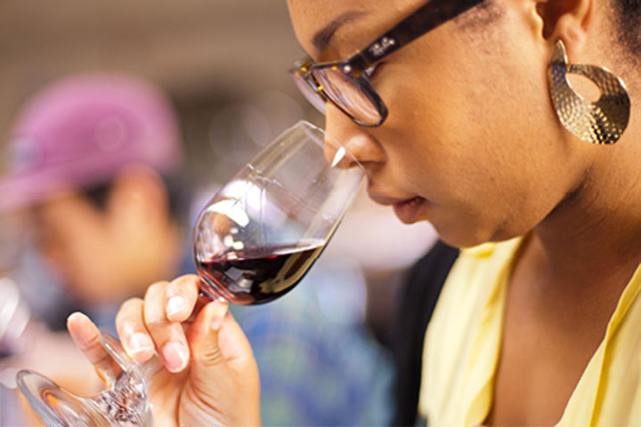 A woman holds a glass to smell red wine, as part of tasting, called "nosing" the wine. She wears a pale yellow top, has her hair tied back to reveal gold earrings and wears dark rimmed glasses.