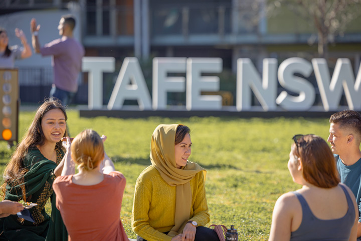 A group of students sit on the grass, talking and relaxing near a large TAFE NSW sign in the background.